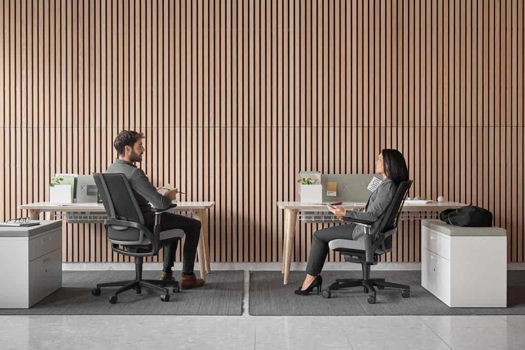 Man and woman wearing smart grey clothes having a conversation sat at desks against a wooden wall
